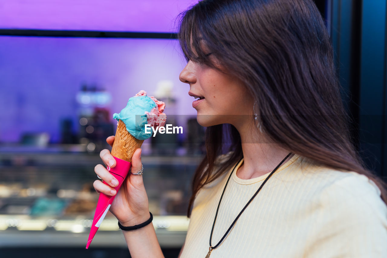 Crop cheerful young female in pendant and earrings with delicious gelato in waffle cone looking away on street