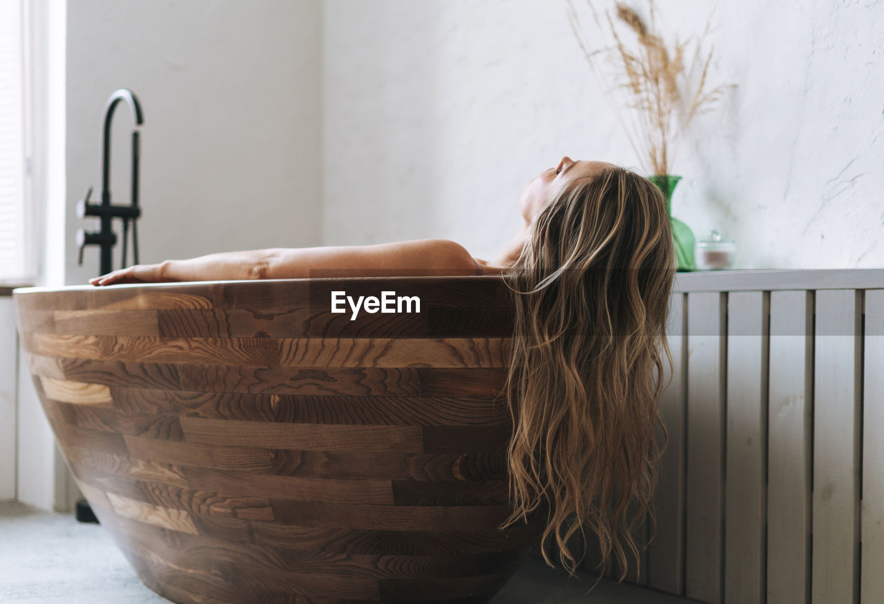 Blonde sensual young woman with long hair takes wooden bath relax at modern bathroom 