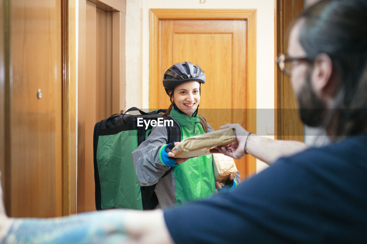 Client receiving paper bag with food from glad delivery woman with thermal backpack through apartment door