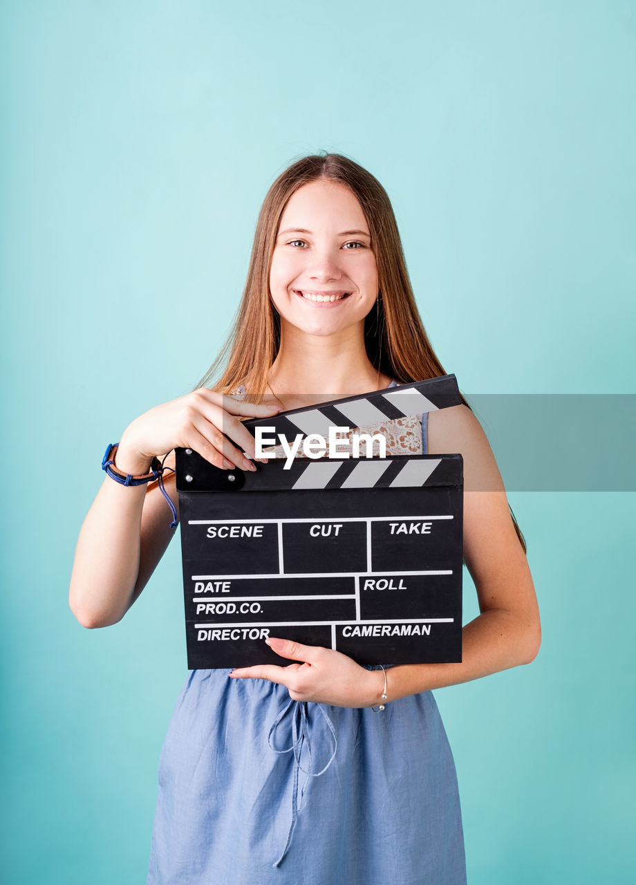 Happy smiling teenager girl in a blue dress holding a clapper board isolated on blue background