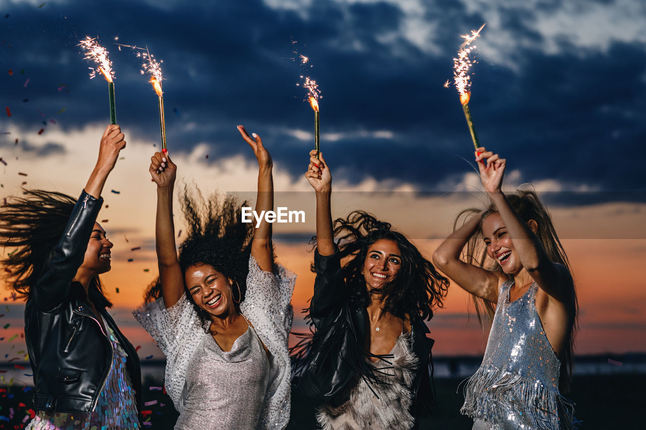 Cheerful friends holding sparklers while standing against sky