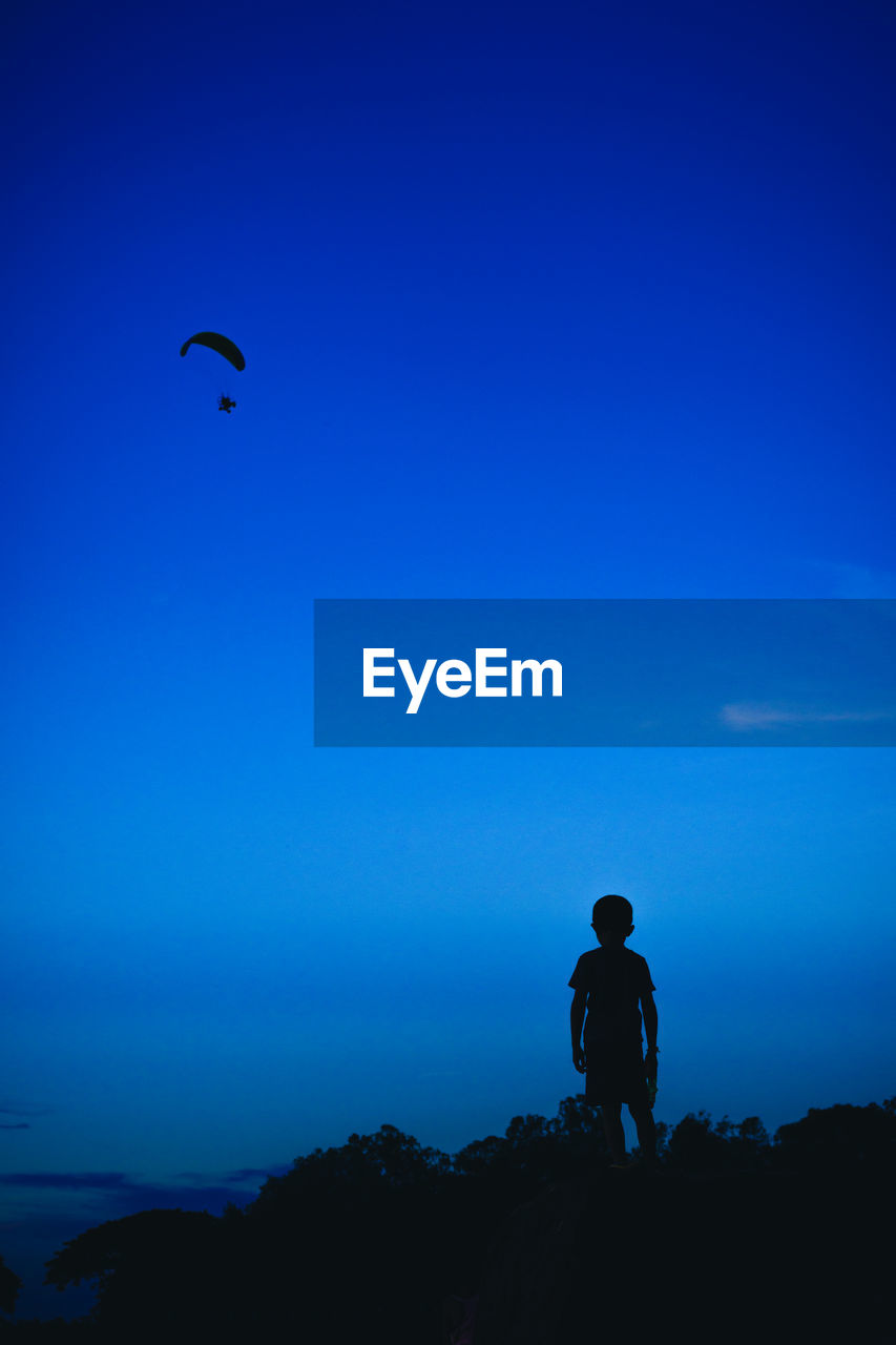 A silhouette boy looking at a blurred silhouette parachute with the dark blue background