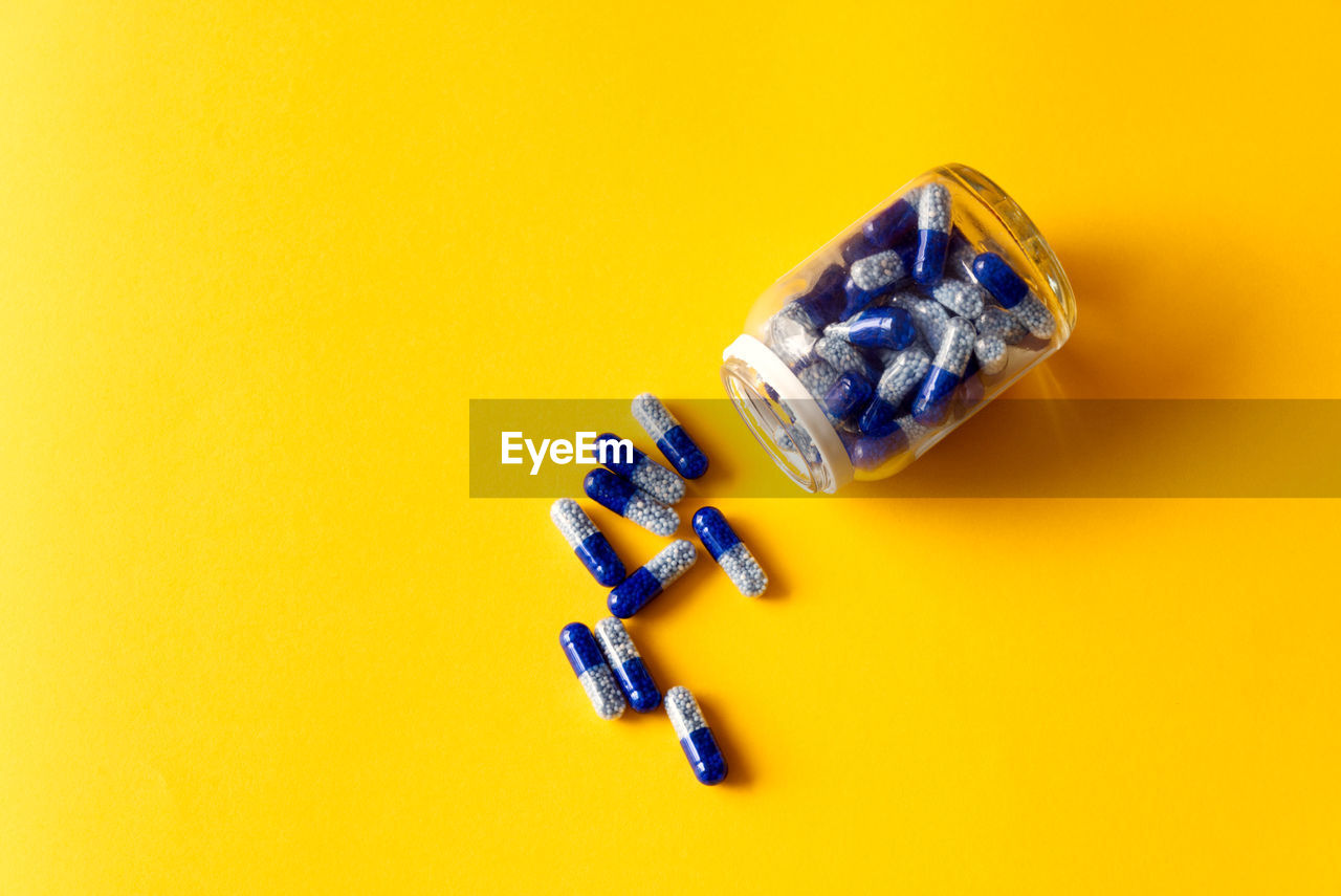 Blue vitamin capsules spilled out of jar on yellow background