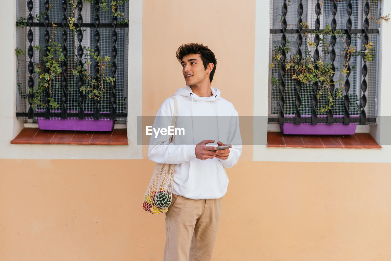 Young male standing on city street with eco friendly mesh bag with groceries and using smartphone while looking away