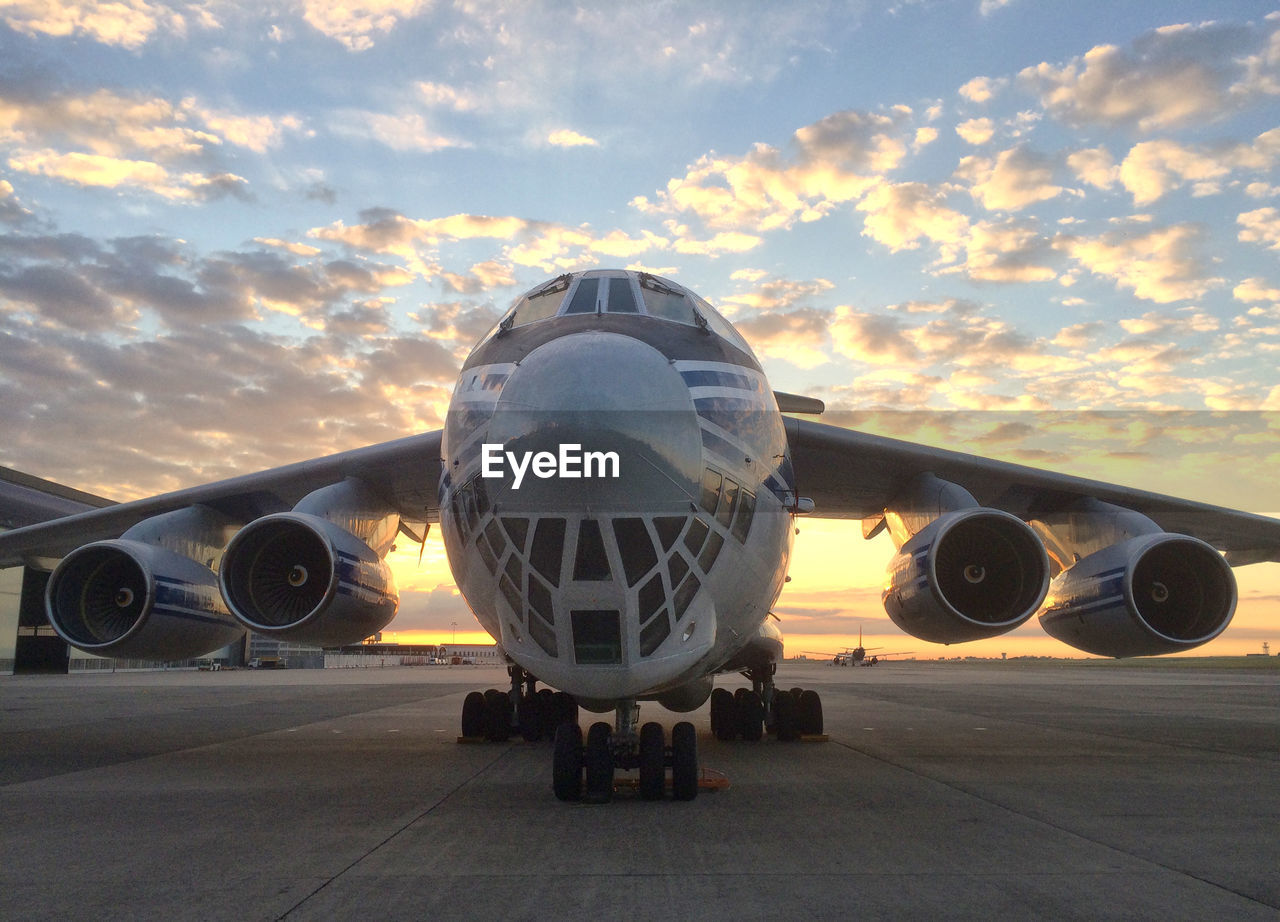 VIEW OF AIRPLANE AT AIRPORT RUNWAY AGAINST SKY DURING SUNSET