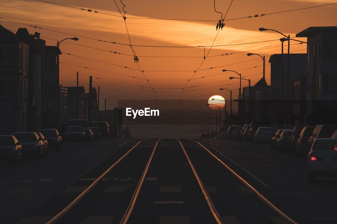 Railroad tracks amidst cars and buildings against sky during sunset