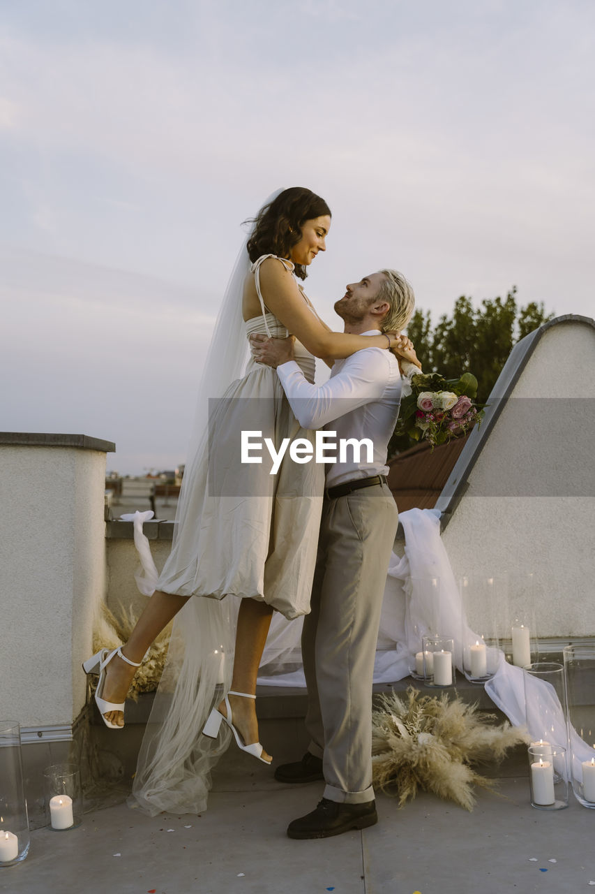 Side view of groom holding bride in arms while standing on rooftop