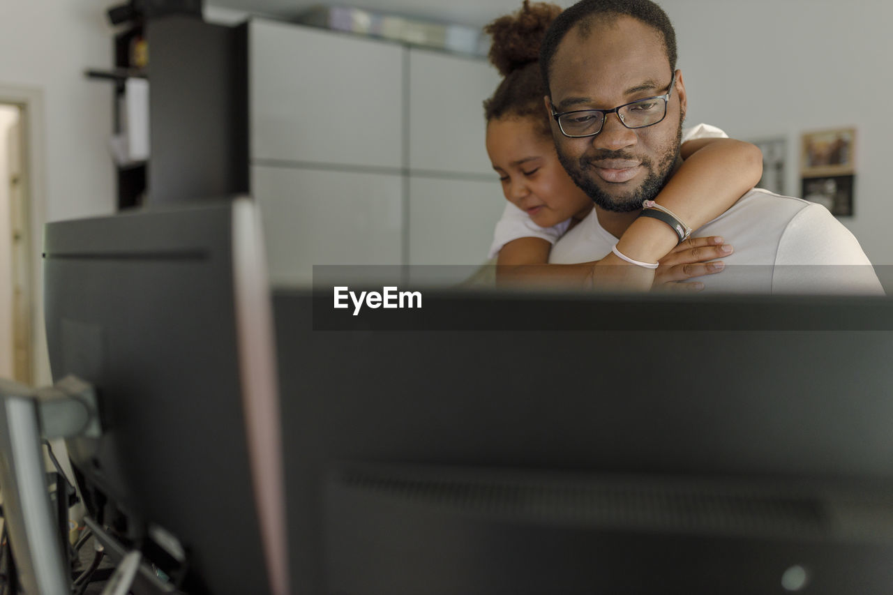 Smiling freelancer working on computer with daughter hugging him from behind at home