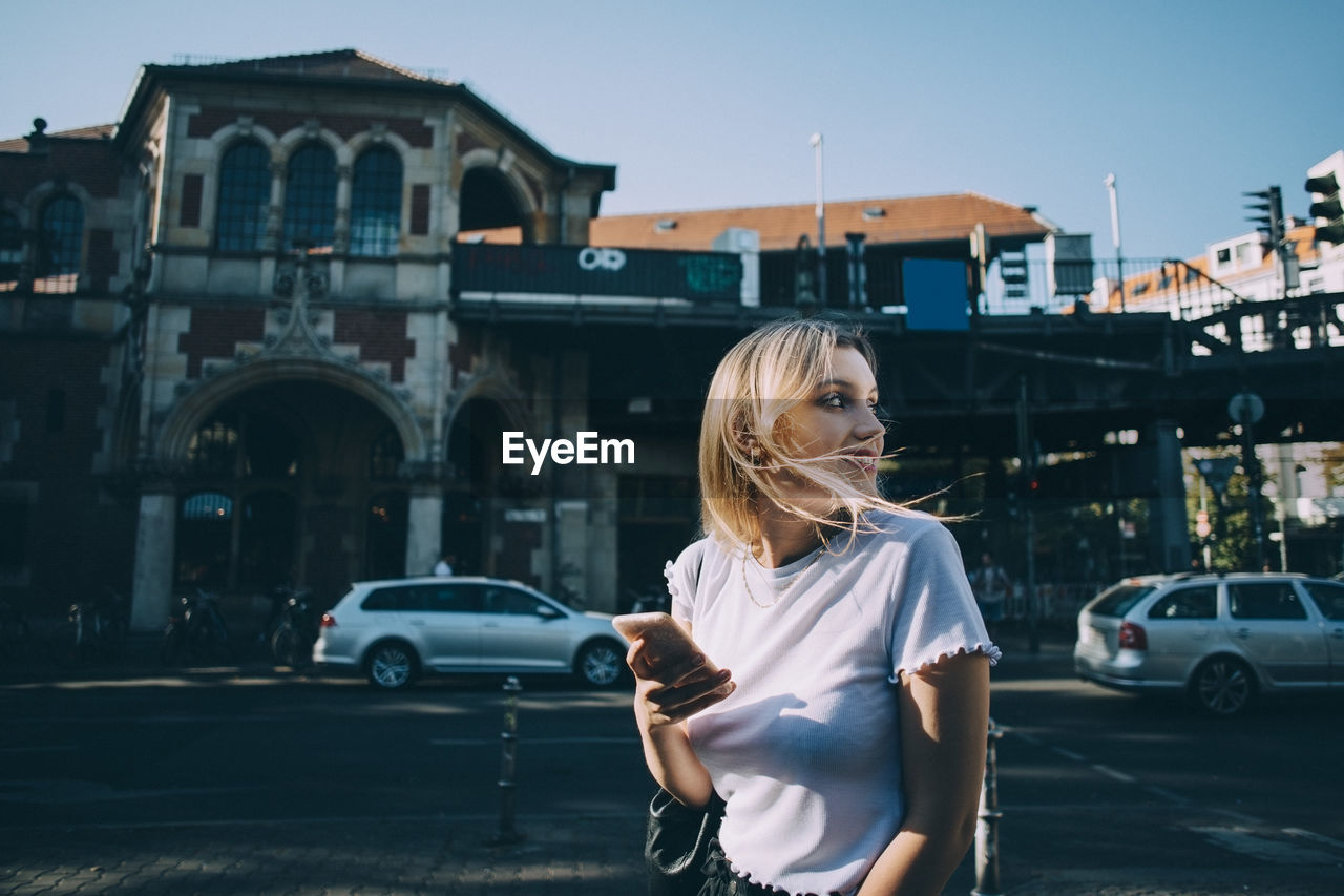 Young woman looking over shoulder while holding mobile phone on street in city