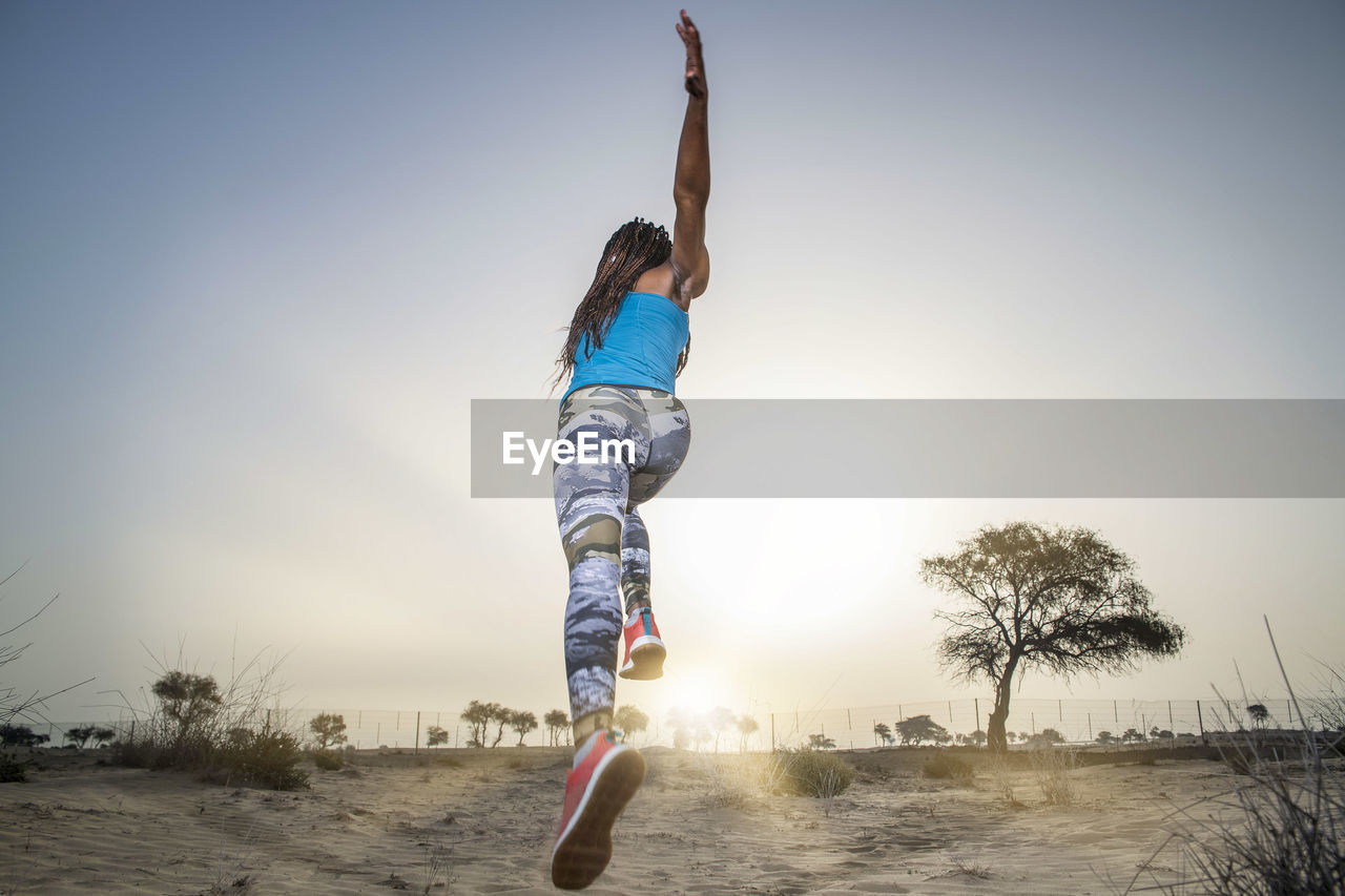 Full length of woman jumping while running in desert against clear sky during sunset