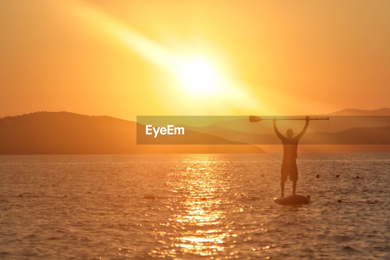 Silhouette man paddleboarding in sea against orange sky