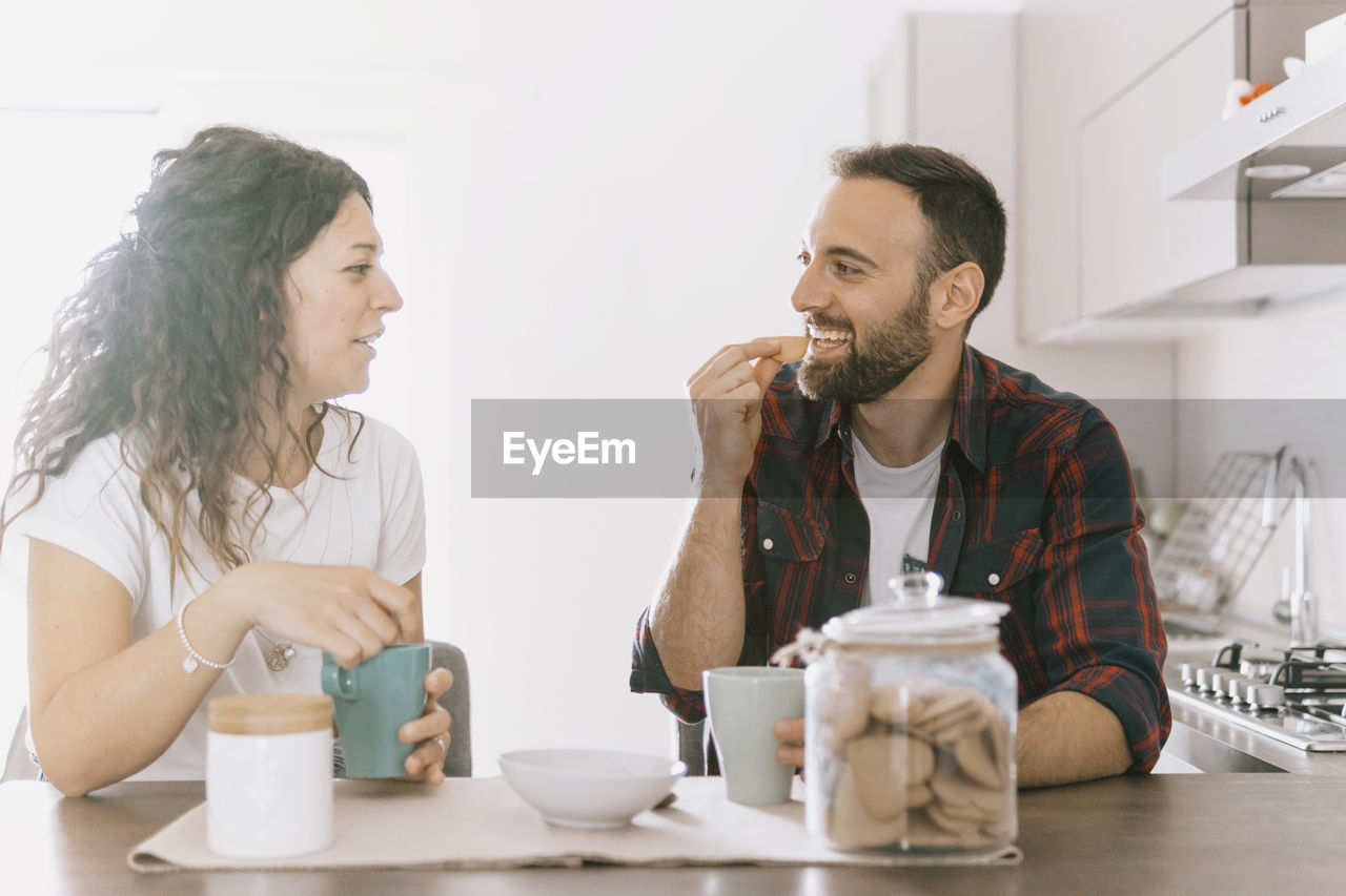 adult, two people, food and drink, indoors, men, person, togetherness, women, drink, cup, lifestyles, mug, coffee, sitting, table, tableware, coffee cup, domestic life, clothing, smiling, young adult, food, female, emotion, happiness, bonding, refreshment, hot drink, casual clothing, domestic room, business, communication, drinking, home interior, talking, enjoyment, human face, positive emotion, friendship, love, tea, home, beard