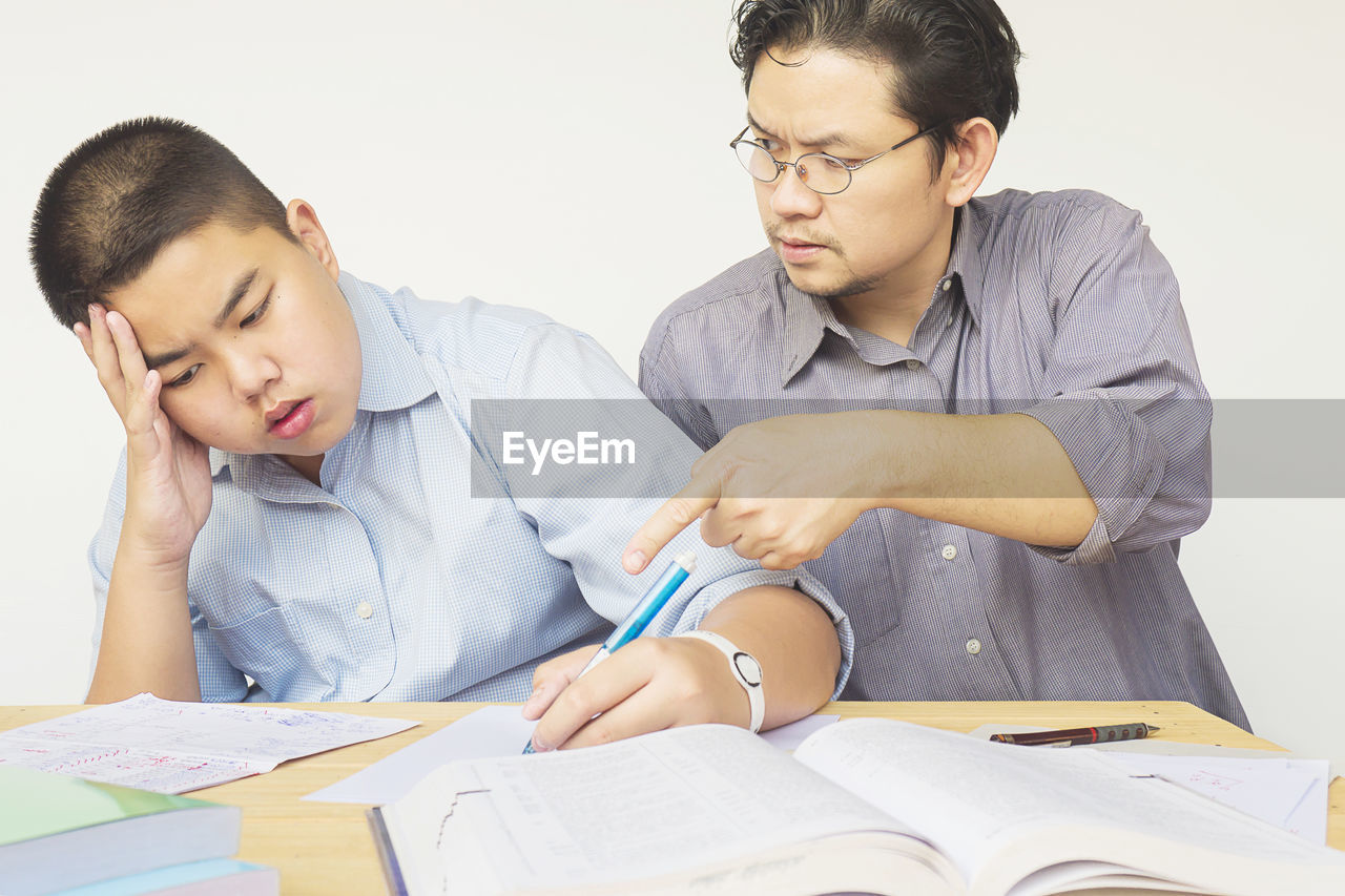 Frustrated father teaching son while sitting at table against white background