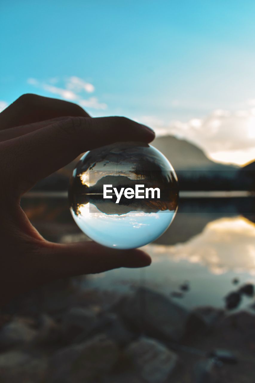 Close-up of hand holding crystal ball against lake and sky during sunset