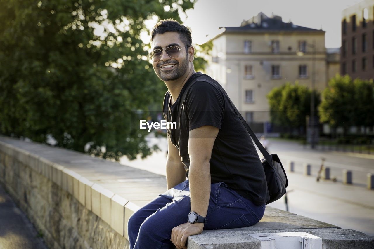 Indian arab or hispanic young man in black shirt with a shoulder strap bag and sunglasses sitting