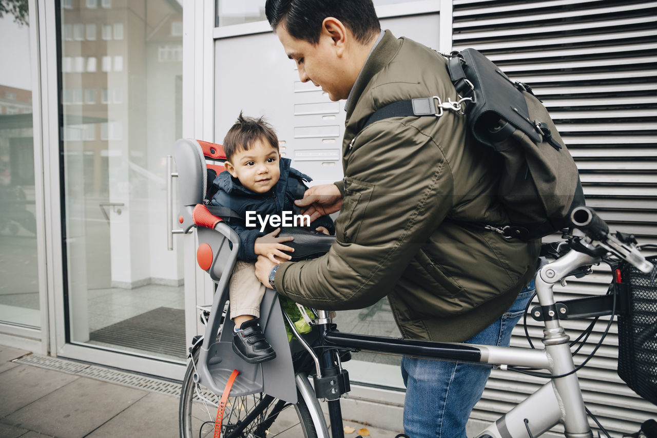Father holding son sitting in safety seat of bicycle on sidewalk in city