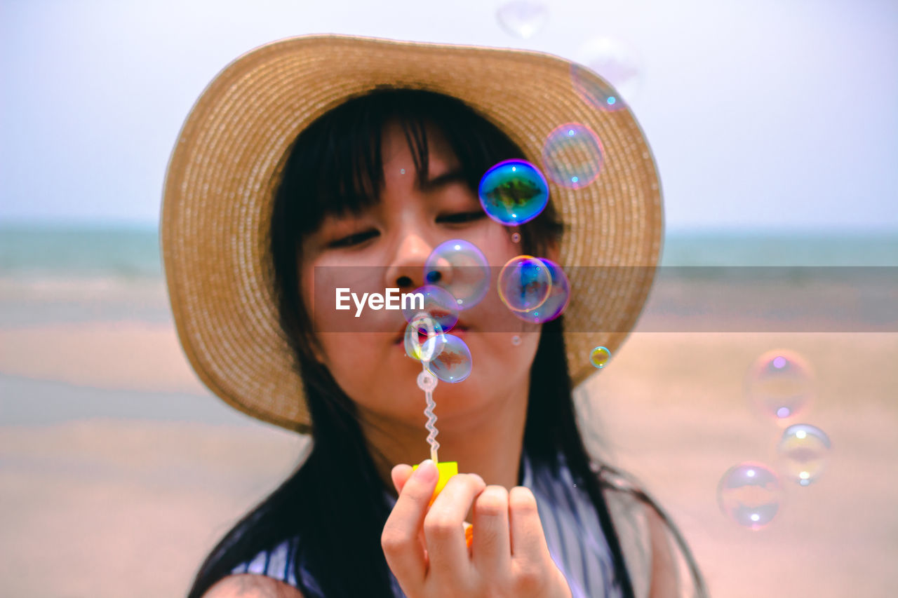 Close-up of young woman wearing hat blowing bubbles while standing against sky