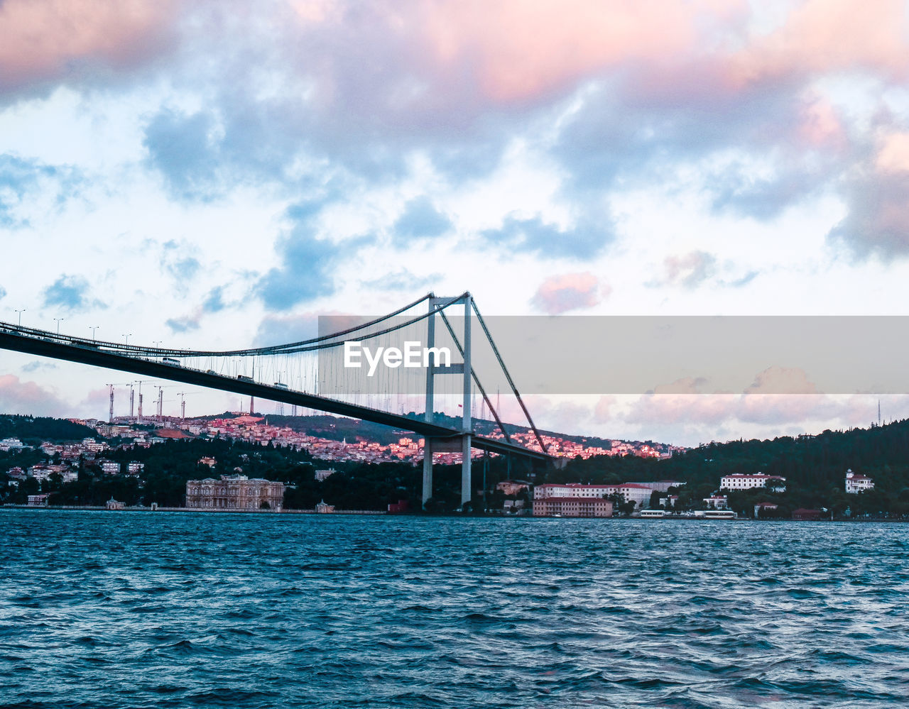 architecture, bridge, water, built structure, transportation, sky, travel destinations, cloud, city, suspension bridge, nature, travel, sea, tourism, engineering, building exterior, bay, bay of water, outdoors, cityscape, mode of transportation, vehicle, beauty in nature, scenics - nature, landscape, city life, nautical vessel, waterfront, cable-stayed bridge, dramatic sky, day, copy space, rippled, building, environment, motion, blue