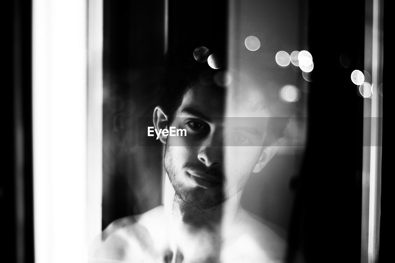 Portrait of shirtless young man looking through window at home