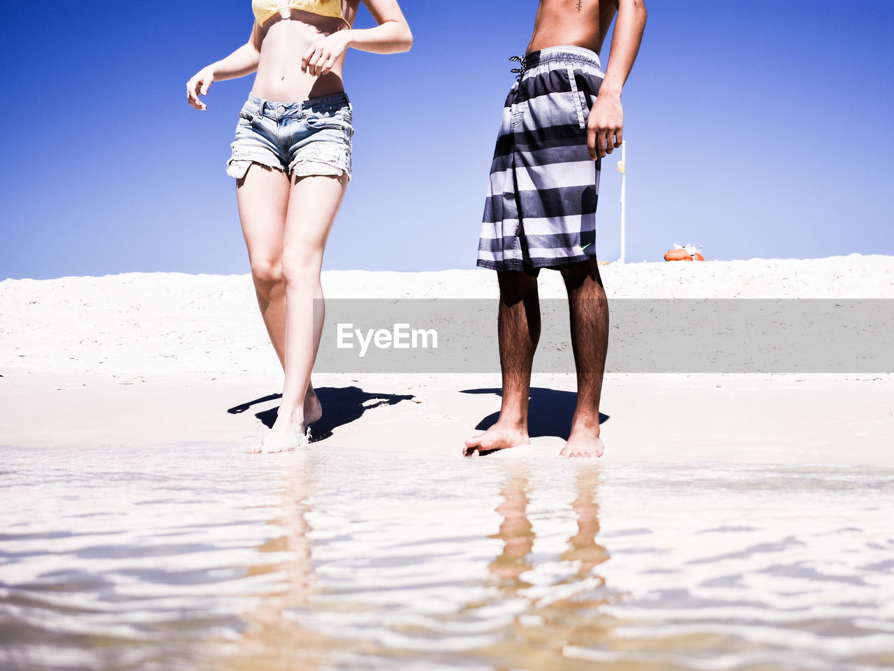 Low section of friends standing on shore at beach against clear sky