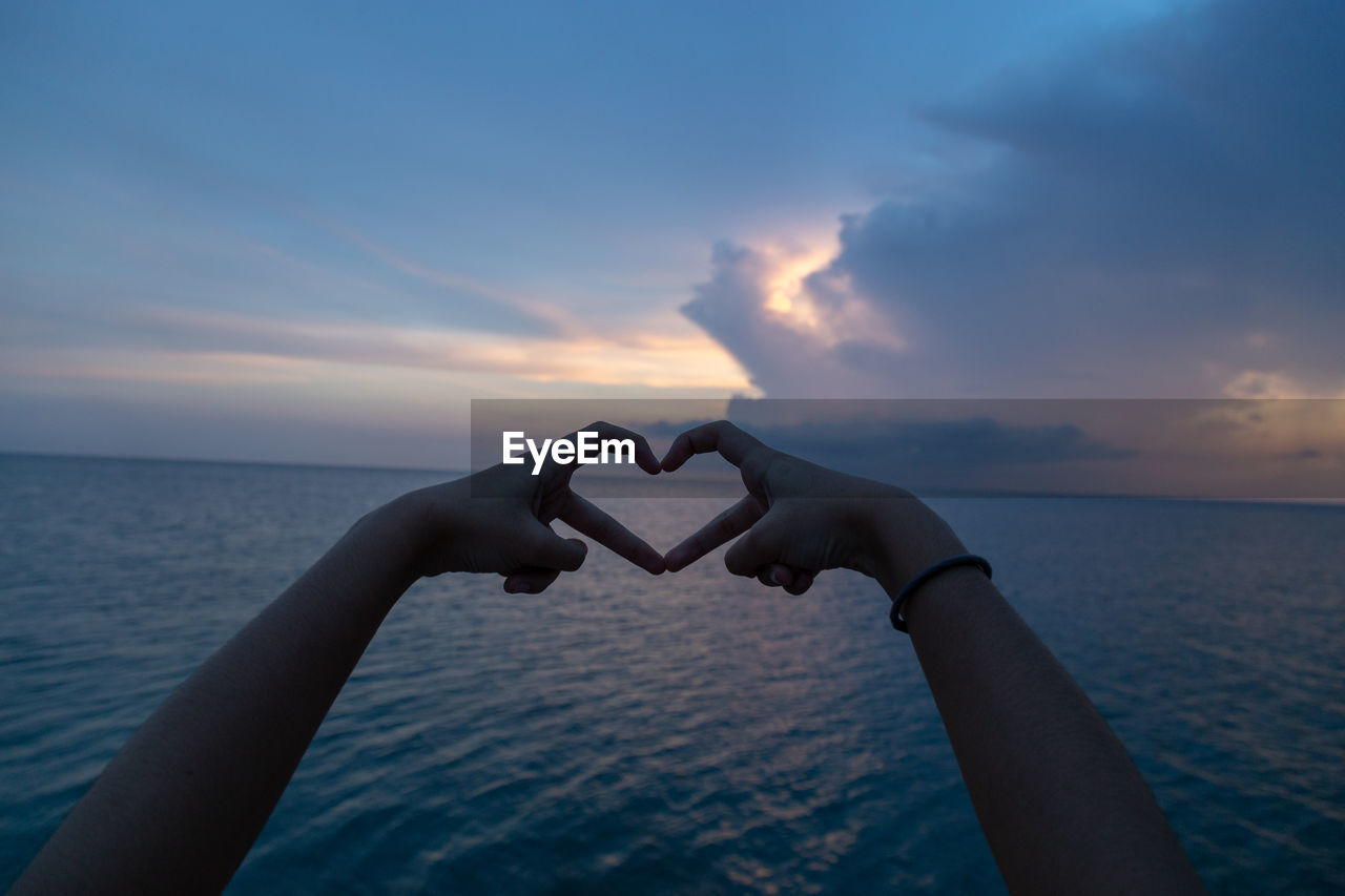 Close-up of hand making heart shape against sea during sunset