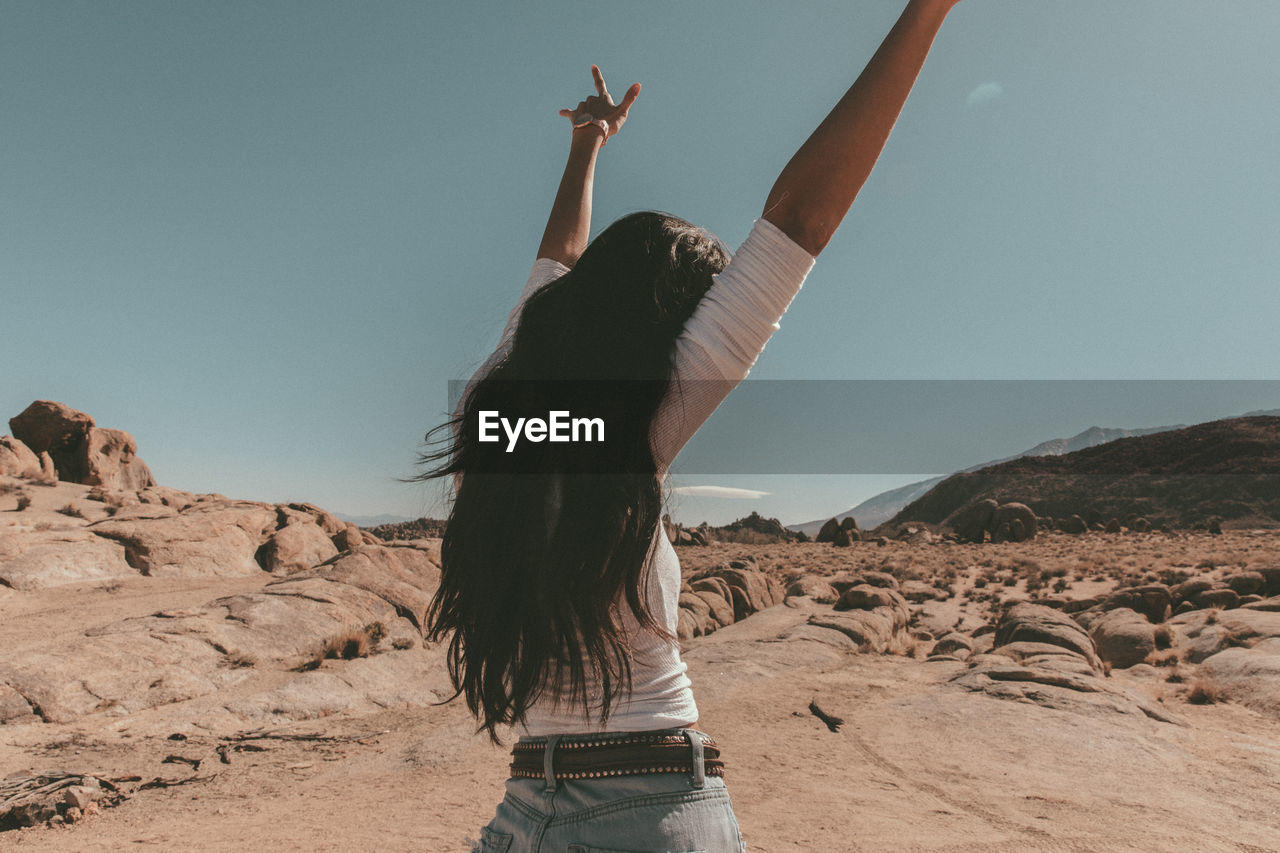 Rear view of young woman with arms raised standing at desert against sky during sunny day