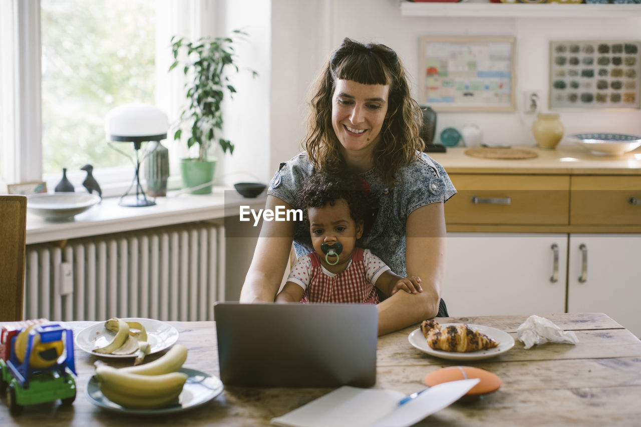 Smiling woman using laptop while sitting with daughter at dining table in house