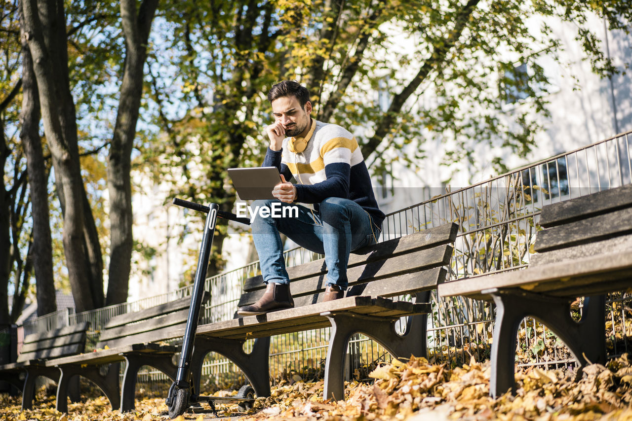 Young man using digital tablet while sitting by electric push scooter on bench at park during sunny day