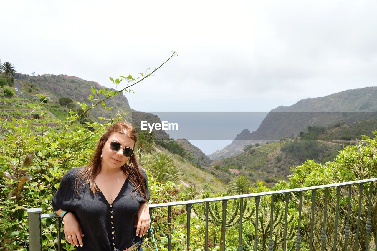 Portrait of young woman standing railing against mountain range