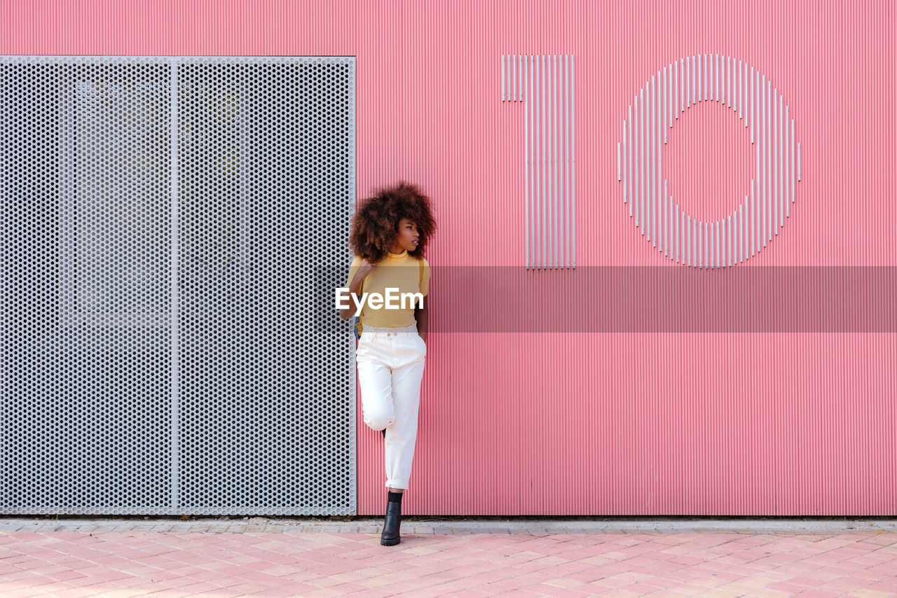 Black woman with afro hair posing in front of a pink wall looking away