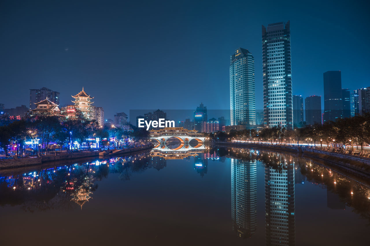 ILLUMINATED BUILDINGS BY RIVER AGAINST SKY