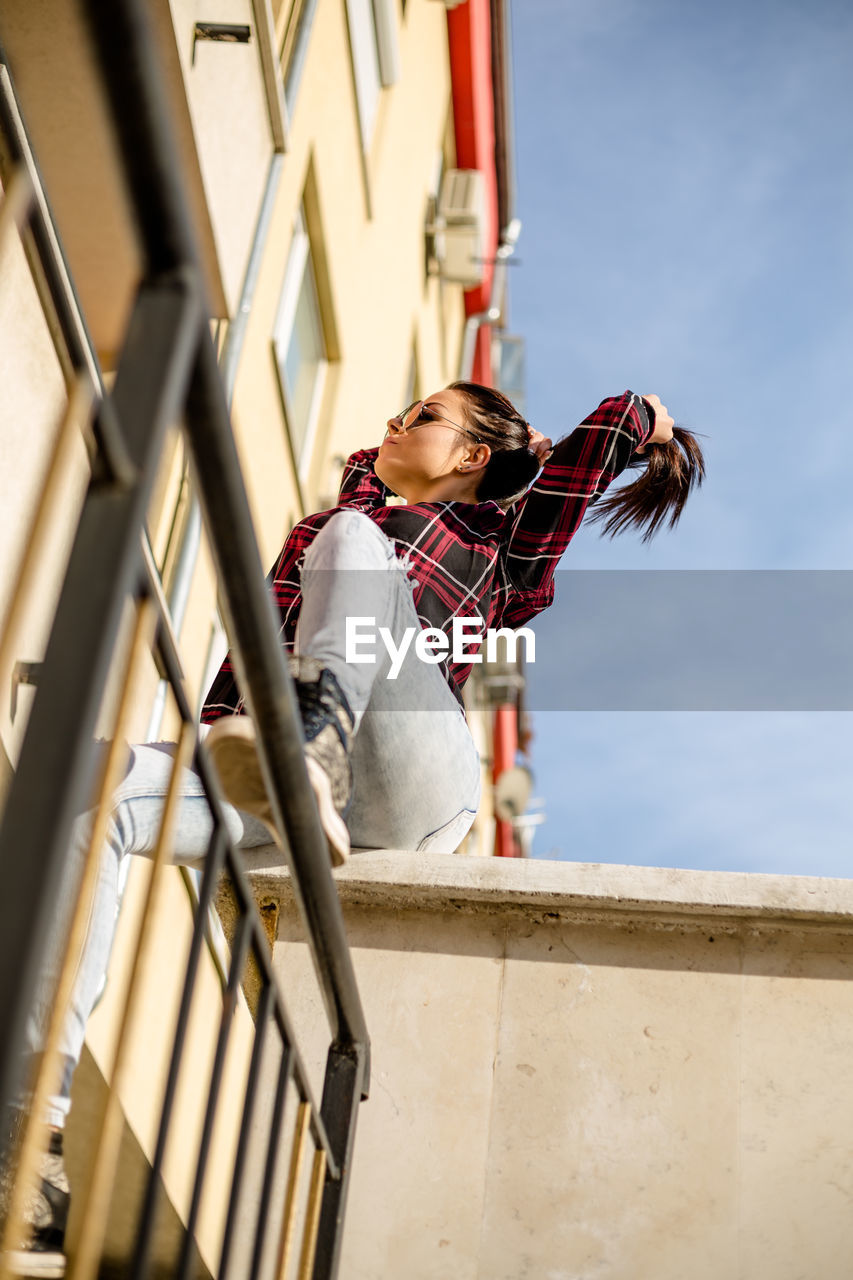Low angle view of woman tying hair while sitting in balcony against sky