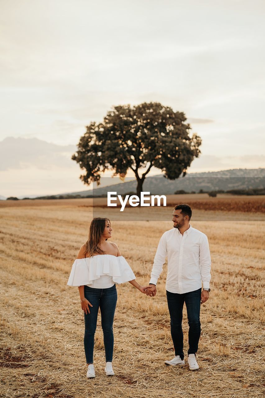 Full body cheerful man and woman holding hands and walking on dry field grass during romantic date in evening in countryside