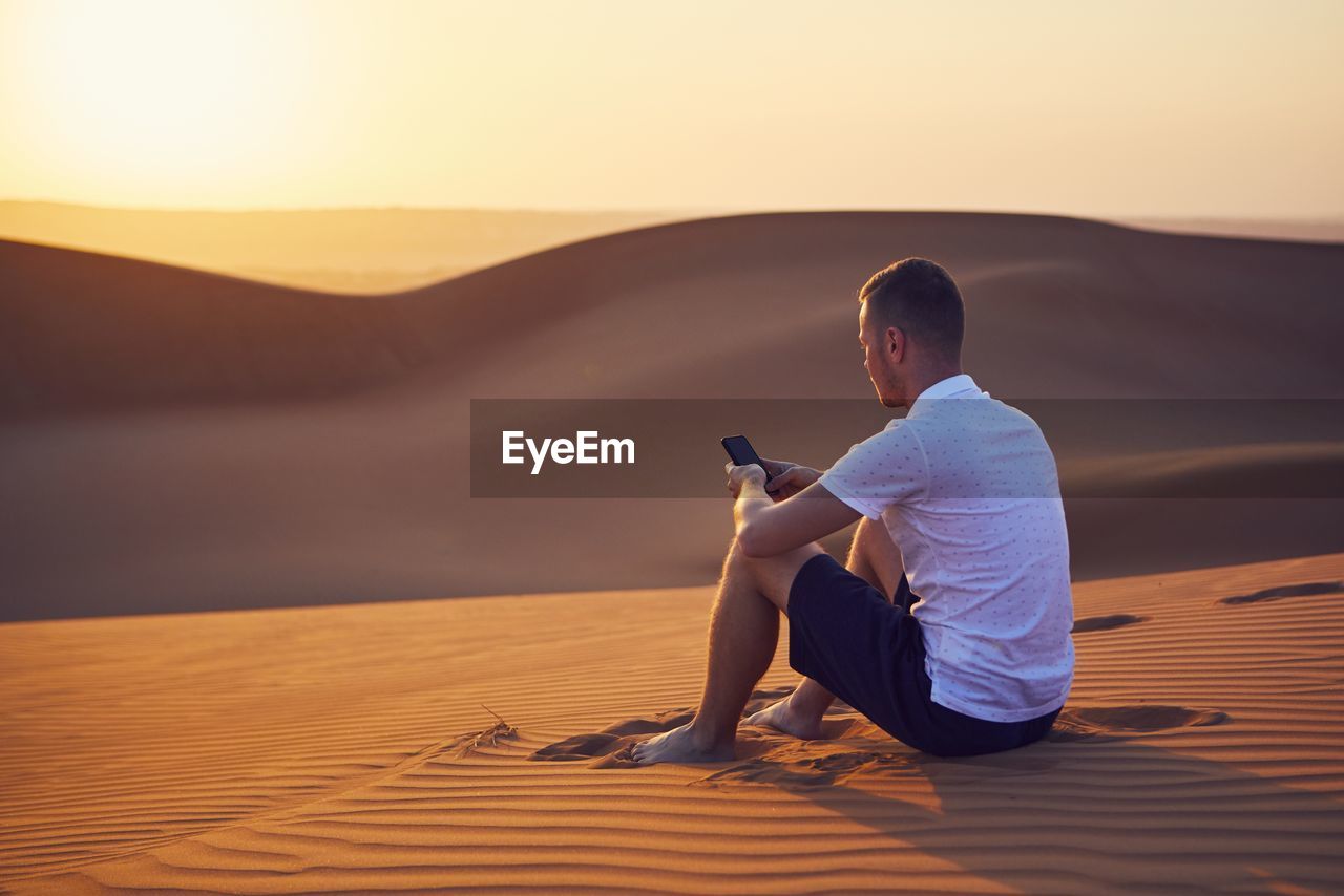 Alone in desert. young man sitting on sand dune and using phone at golden sunset.