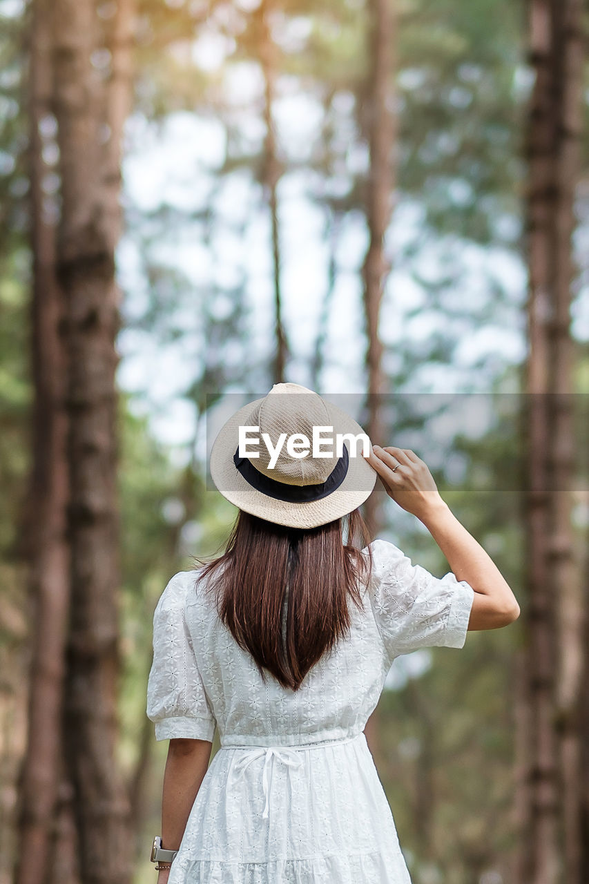 REAR VIEW OF WOMAN WEARING HAT STANDING AGAINST TREES