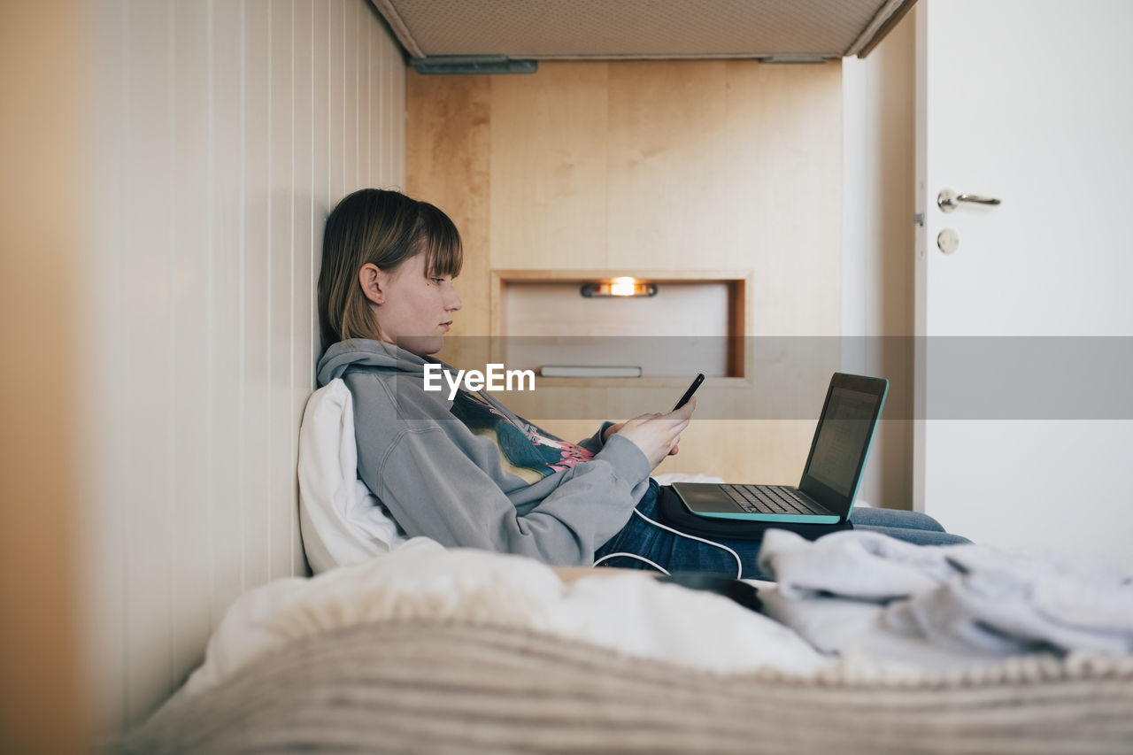 Teenage girl sitting with laptop using smart phone in vacation home