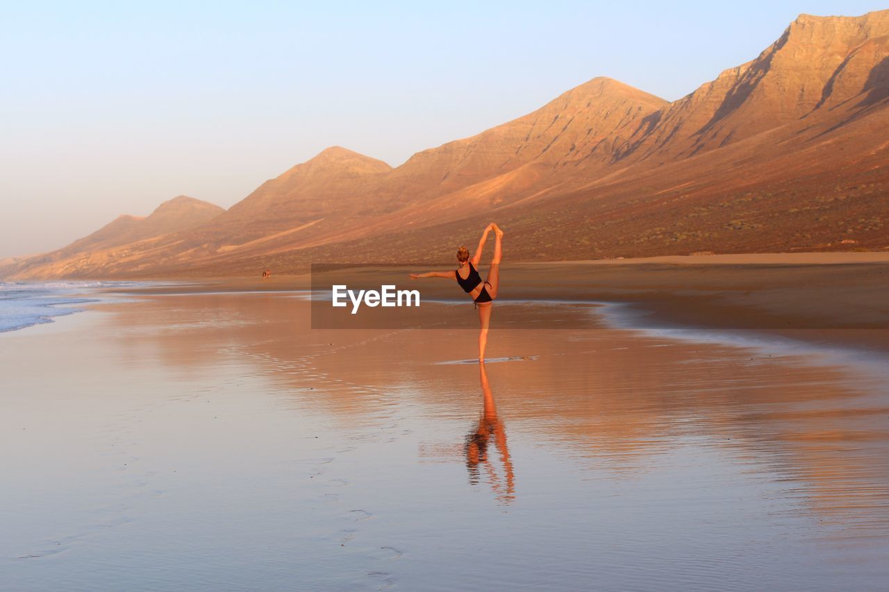 Woman performing yoga while standing at beach against sky