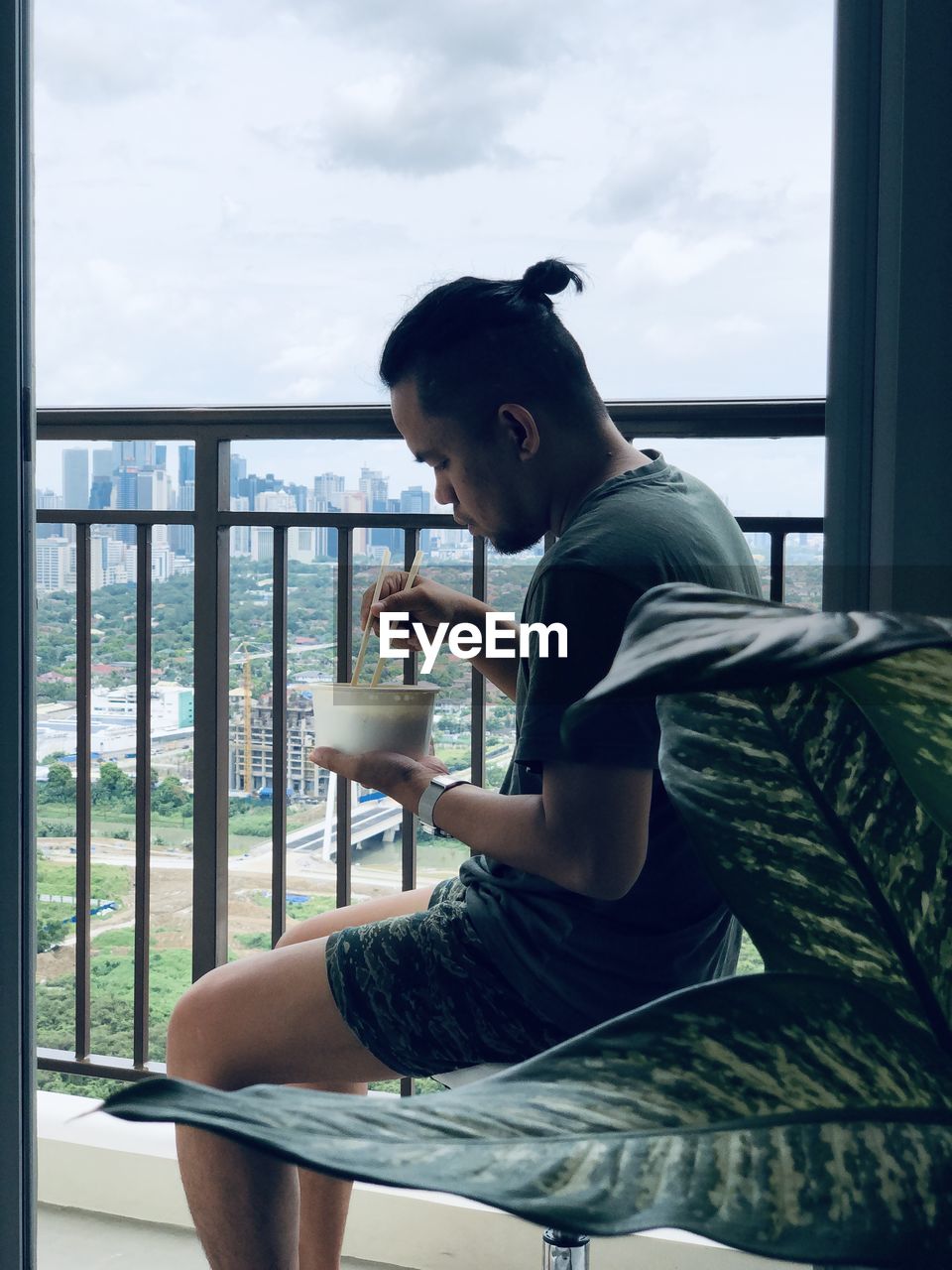 Side view of young man sitting on chair at home against city skyline