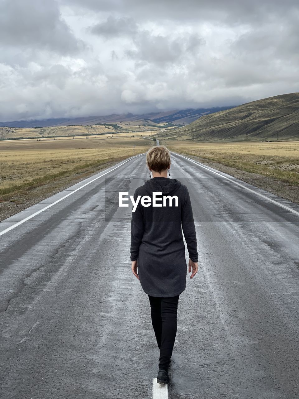 A young woman in a long hooded sweatshirt walking away into the distance on a cloudy fall day. 