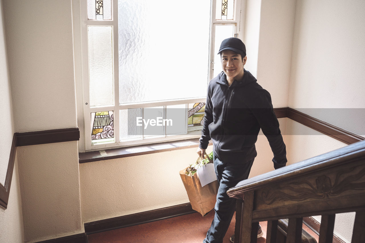 High angle portrait of smiling delivery man with package against window