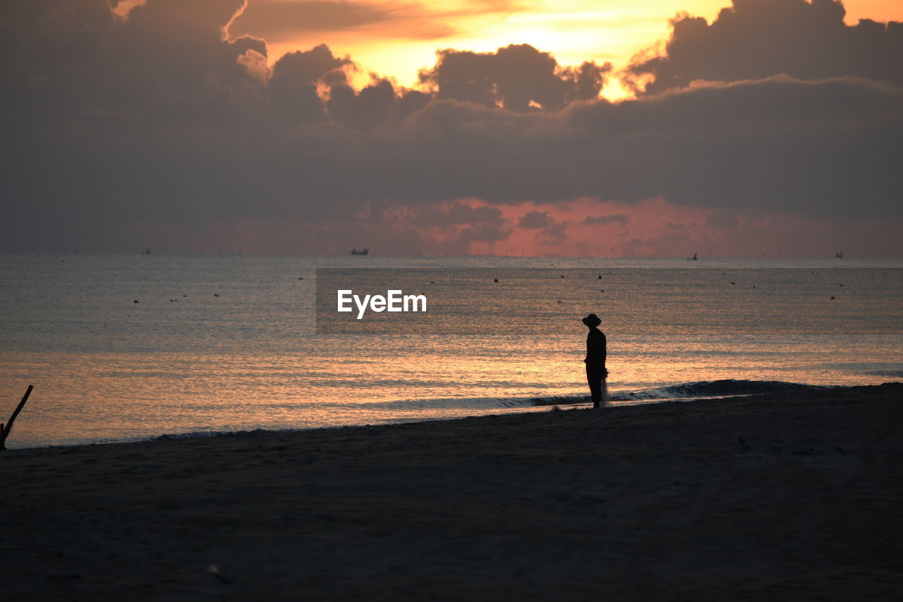 SILHOUETTE MAN STANDING ON BEACH AGAINST ORANGE SKY