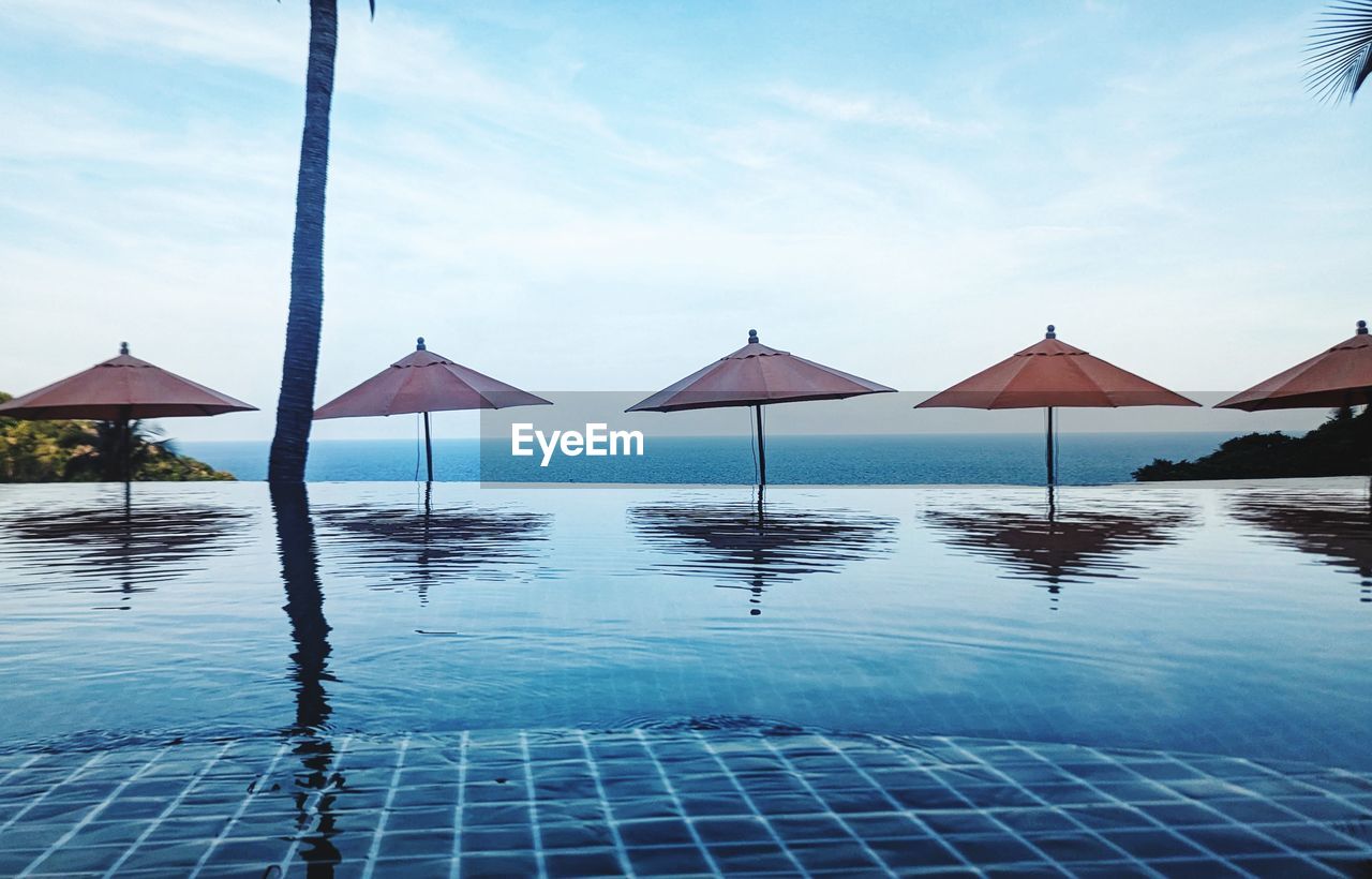 water, sky, nature, sea, tranquility, beauty in nature, tranquil scene, land, ocean, swimming pool, parasol, blue, tropical climate, travel destinations, trip, relaxation, vacation, scenics - nature, umbrella, holiday, idyllic, roof, no people, beach, sunshade, cloud, luxury, day, tourist resort, wealth, outdoors, summer, thatched roof, architecture, travel, lounge chair, infinity pool, chair, beach umbrella, horizon, reflection, horizon over water, environment, sunlight, shade, palm tree, luxury hotel, bay, absence