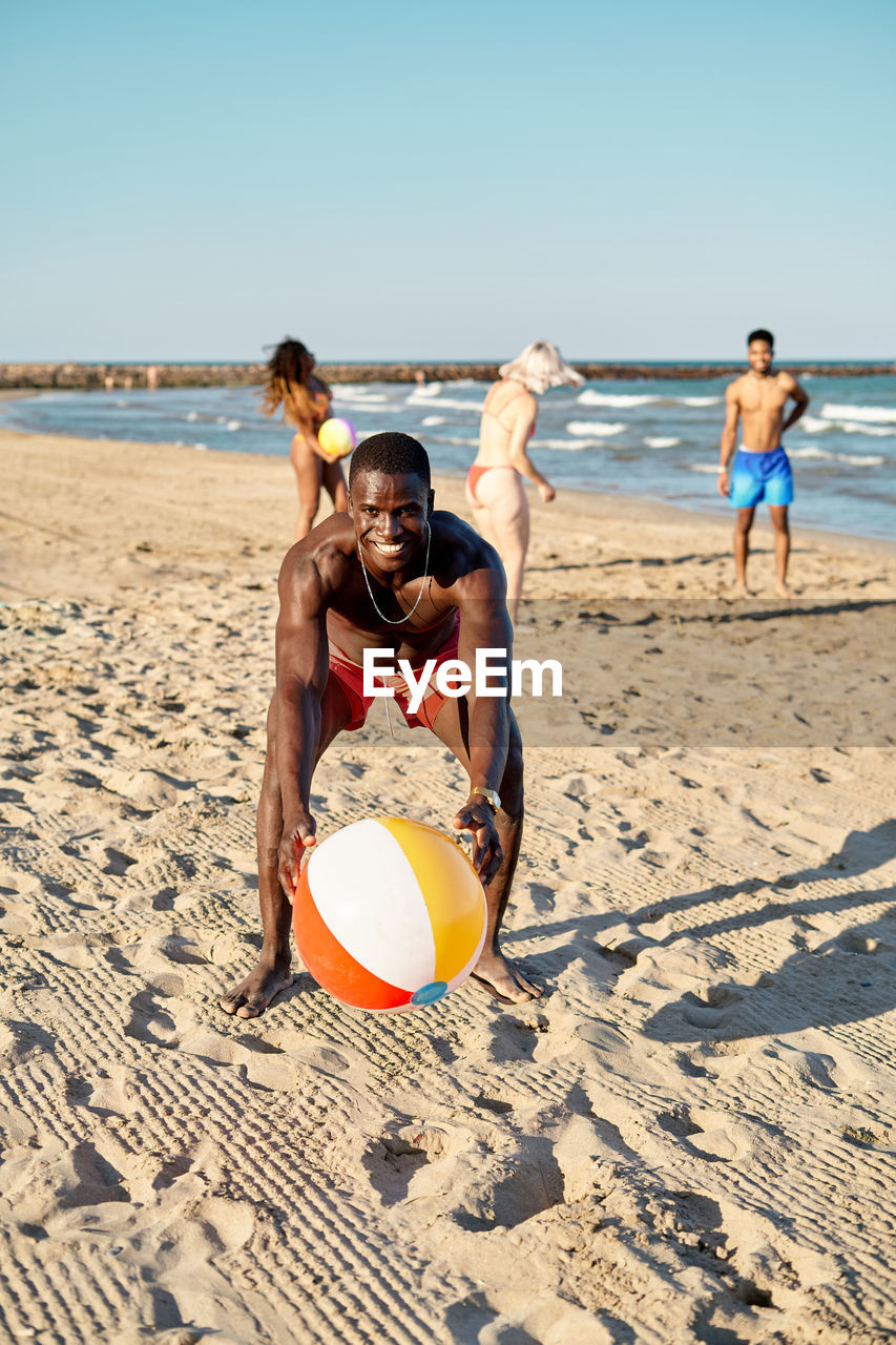 Happy african american male in shorts looking at camera with smile and taking ball from sand while spending time with friend on beach near sea