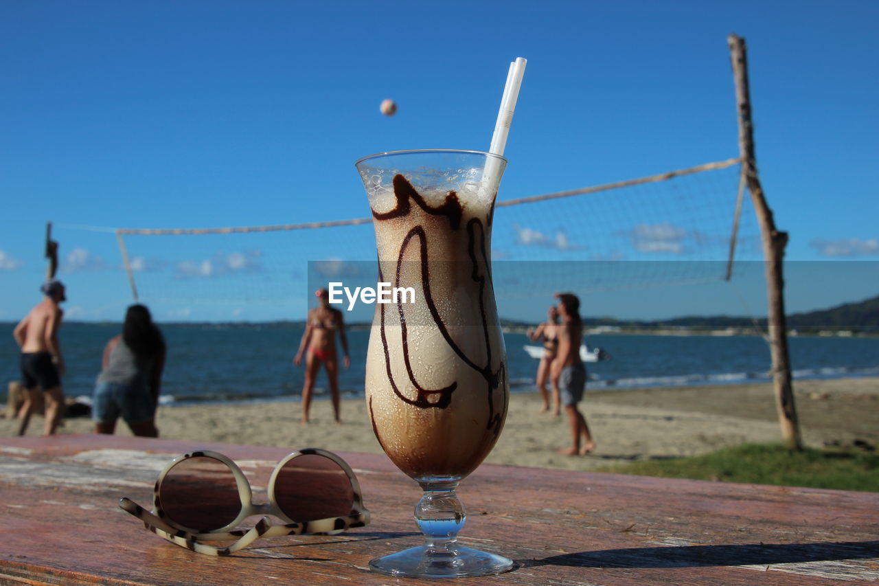 Low angle view of drink and of  of people at beach playing beach volleyball against blue sky