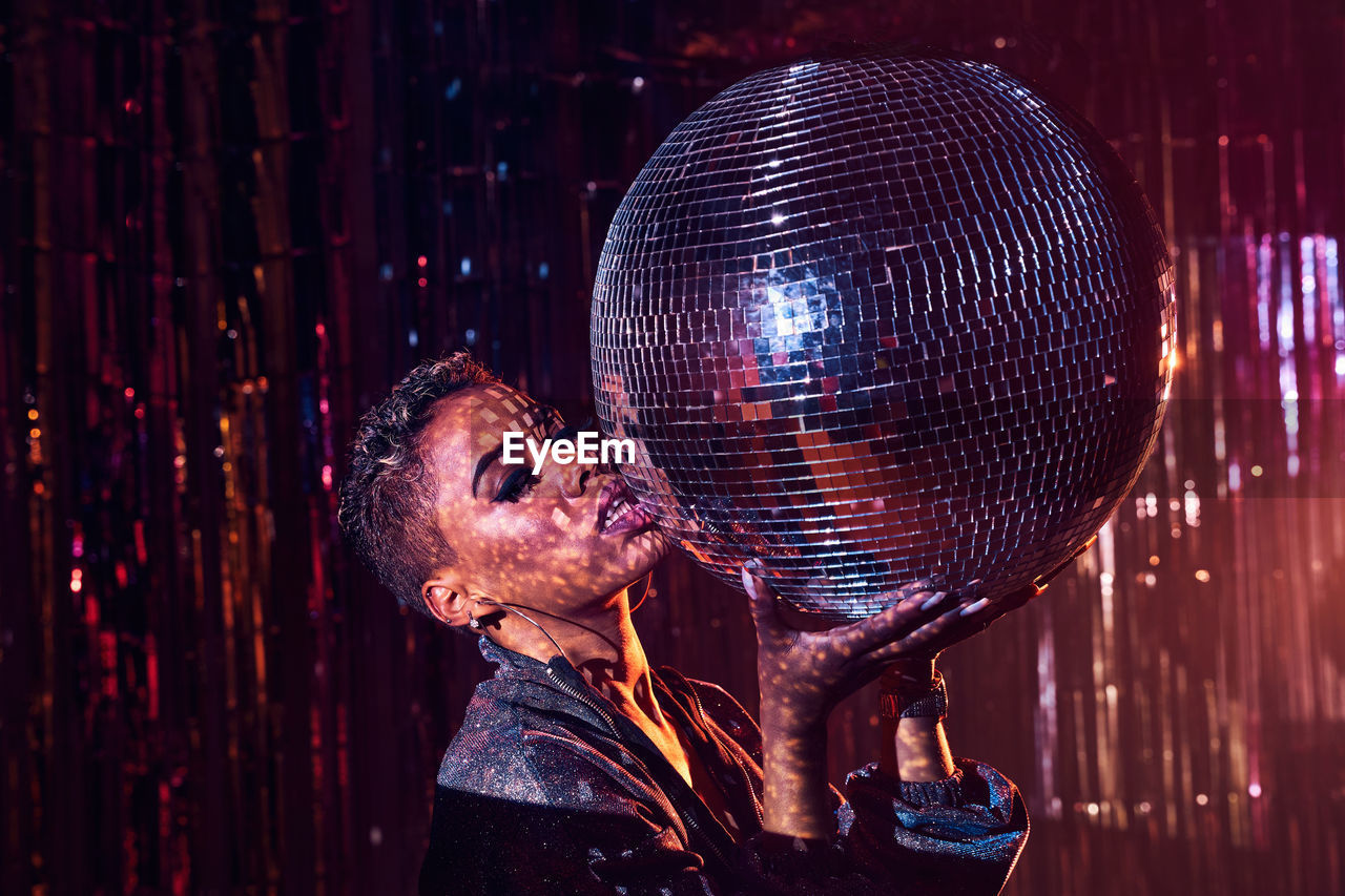 Trendy young ethnic female touching lips while looking at camera and leaning on mirror ball in nightclub