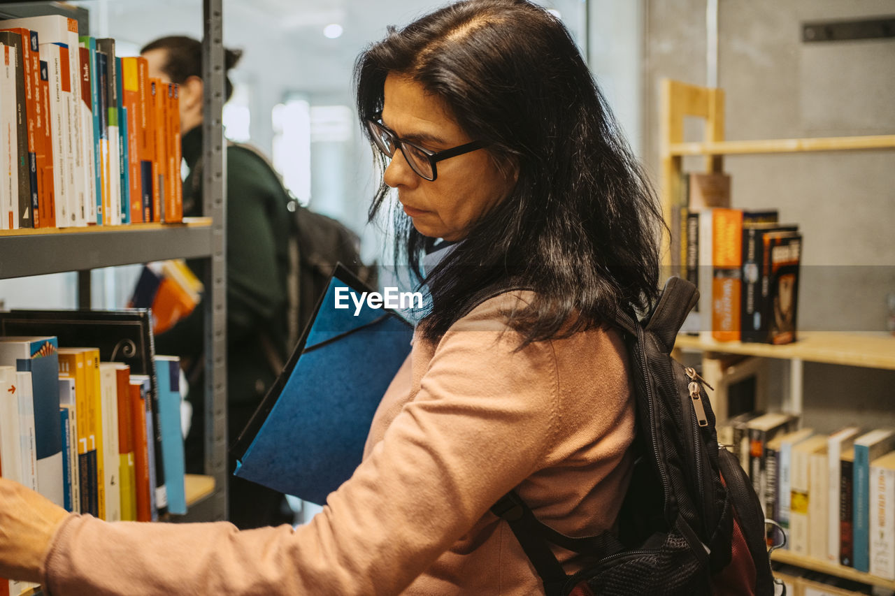 Side view of mature female student searching book on rack in college library