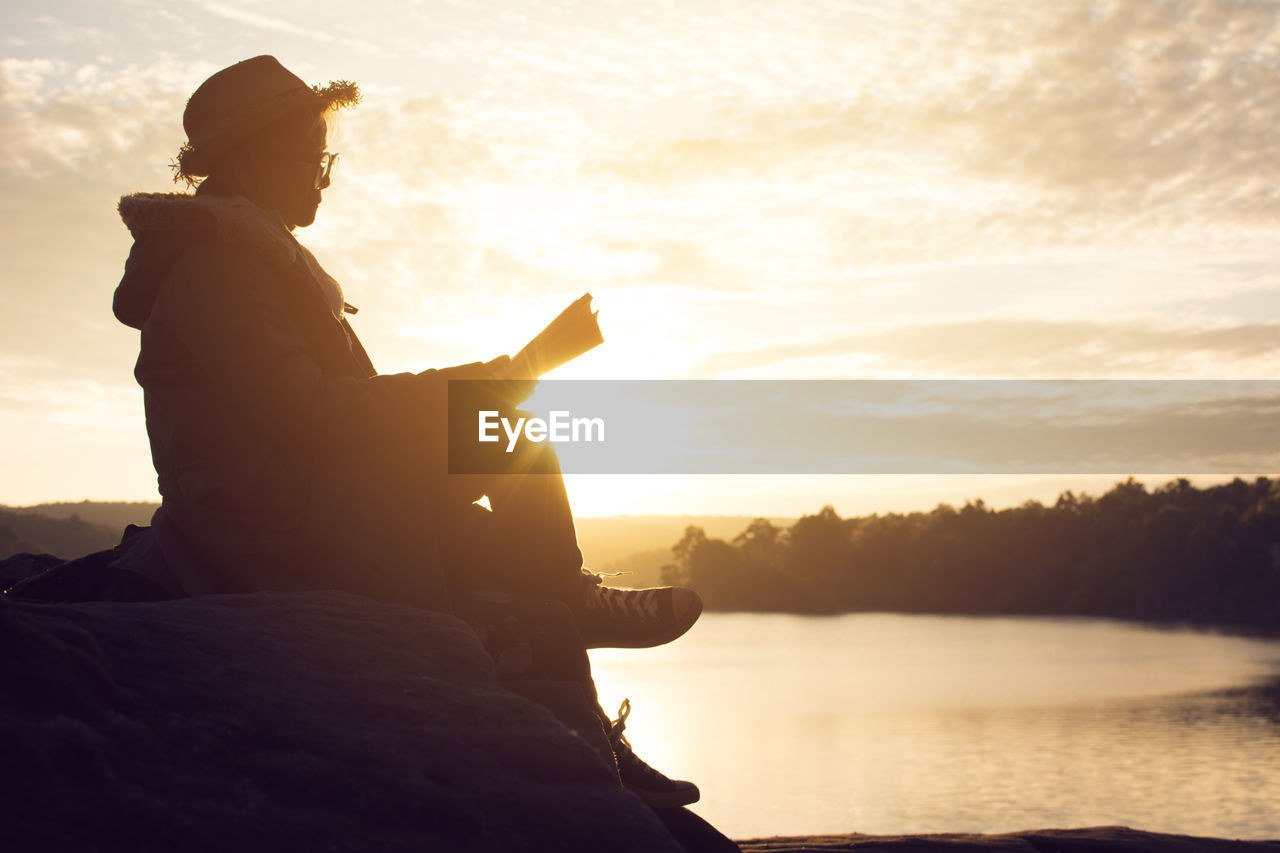 Senior woman sitting on rock by lake against sky