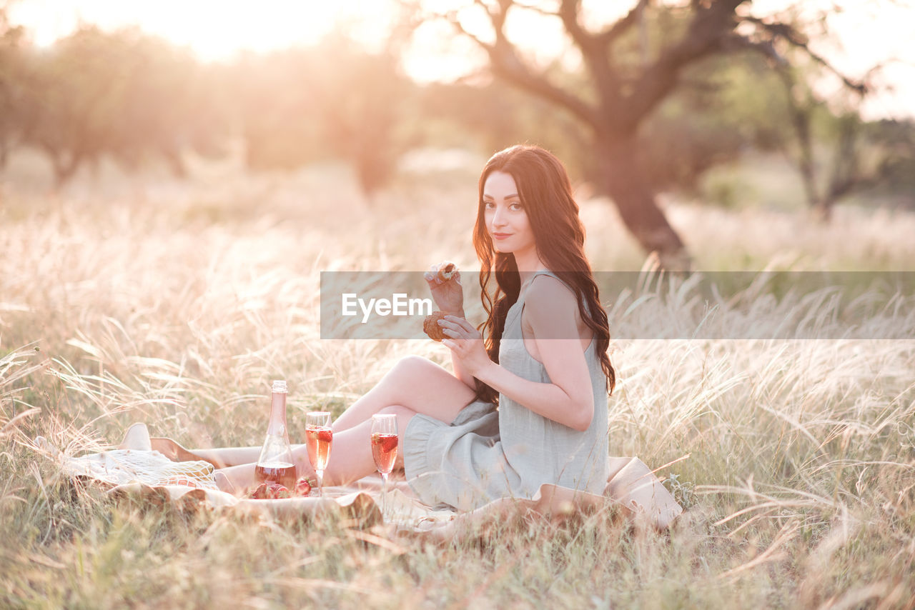 Happy young woman having picnic in meadow with glass of wine, fruit over sunset outdoors. 