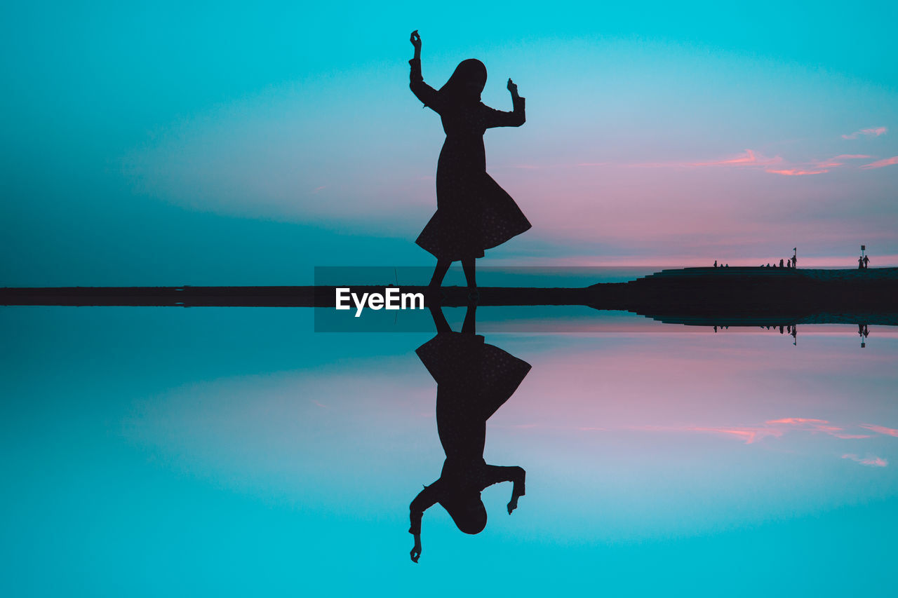 Symmetric image of woman standing by calm lake against sky during sunset