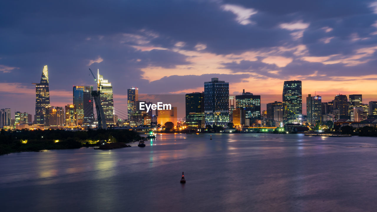 Illuminated buildings by river against sky